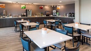 A modern restaurant with white tables and blue chairs, a kitchen area with appliances, and a counter with food items.