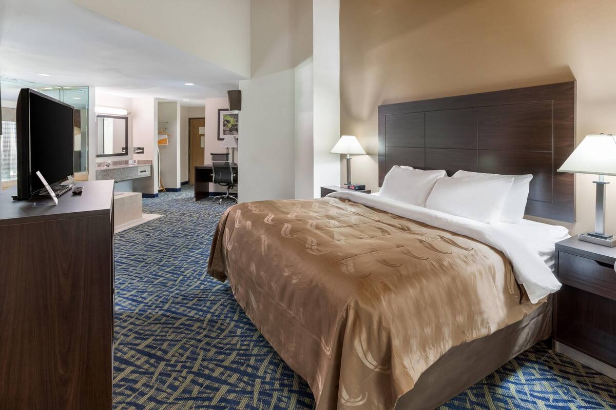 Una habitación de hotel con una cama grande, lámparas de noche, un escritorio con televisor y un baño con espejo y toallero.
