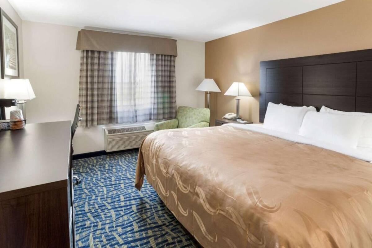 Habitación de hotel con una cama grande, dos lámparas de noche, un escritorio con lámpara, una silla, una ventana con cortinas y un equipo de aire acondicionado.