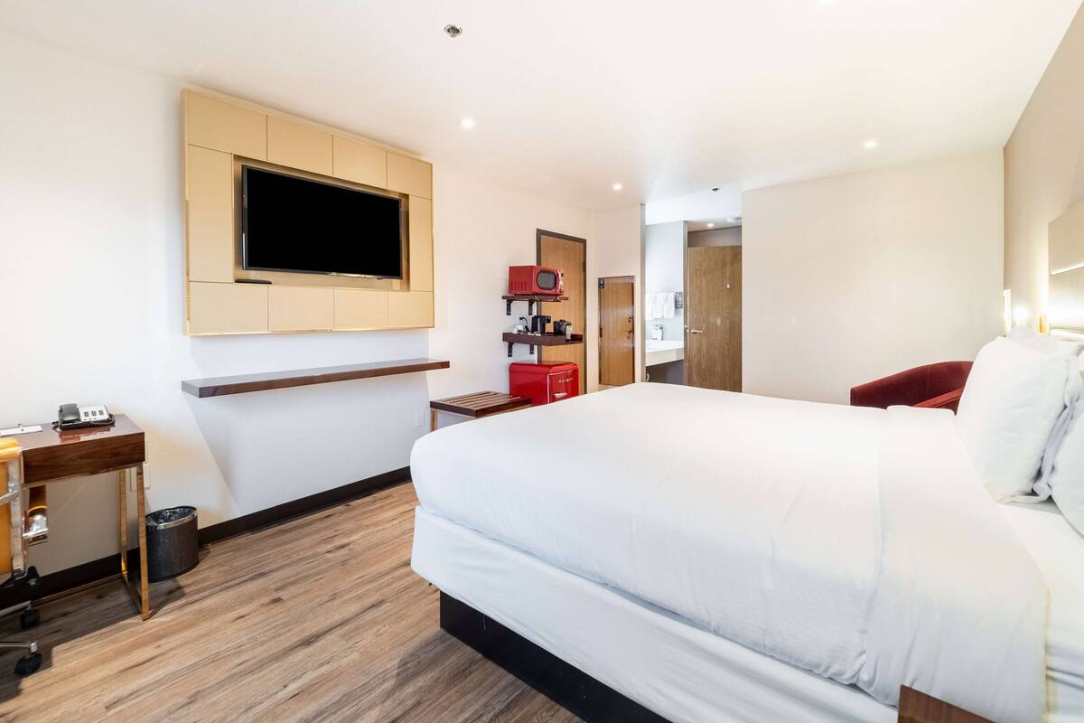 A modern hotel room with a large bed, a desk with a telephone, a TV mounted on the wall, and a small shelf with a radio and a coffee maker.