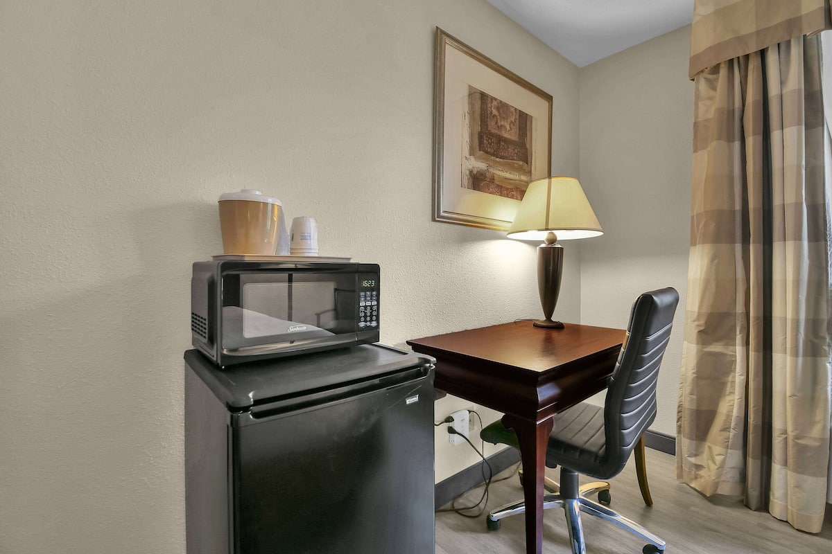 A compact kitchenette with a microwave and refrigerator, a small table with a lamp, a chair, and a framed picture on the wall.