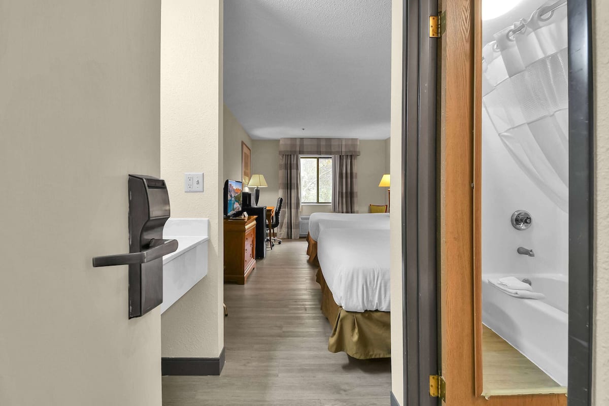 A hotel room with a bed, a desk with a computer, a chair, a window with curtains, and a bathroom with a bathtub.
