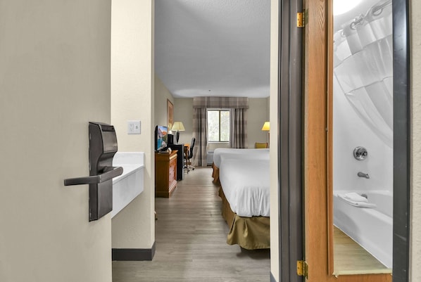 A hotel room with a bed, a desk with a computer, a chair, a window with curtains, and a bathroom with a bathtub.