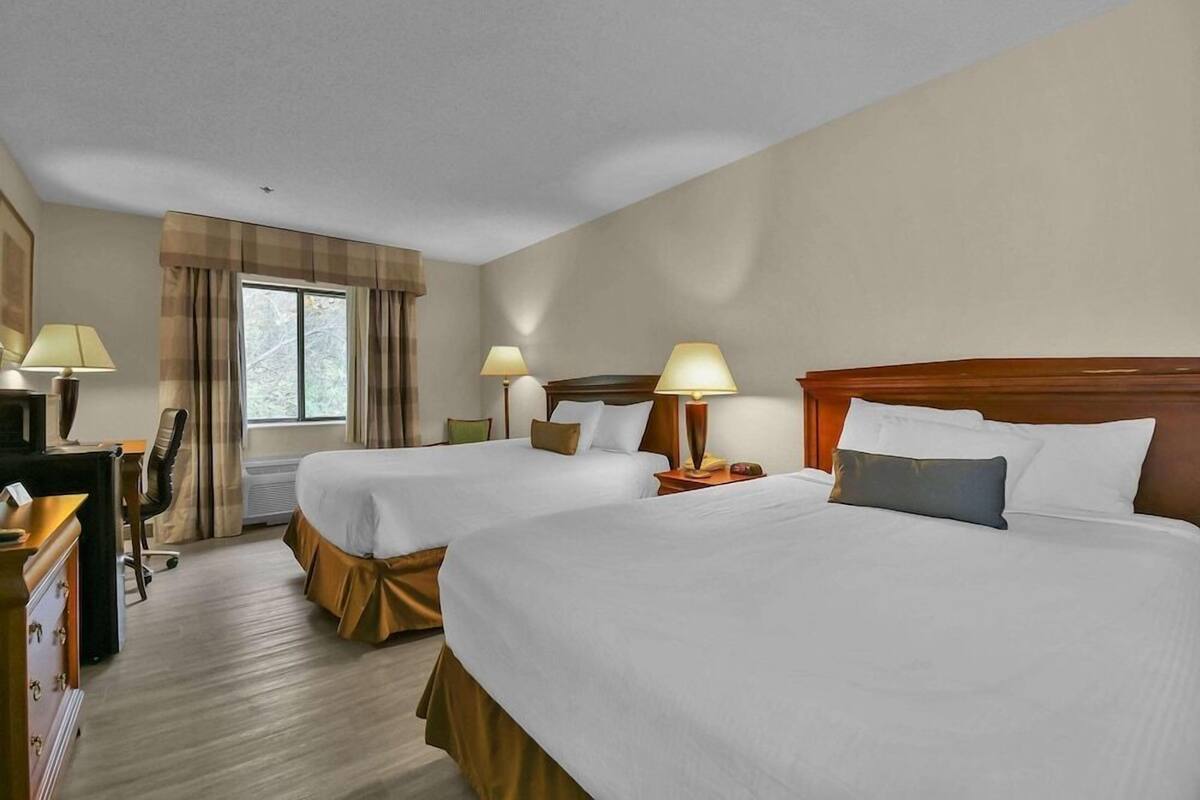 A hotel room with two beds, a desk, a chair, a lamp, and a window with curtains.