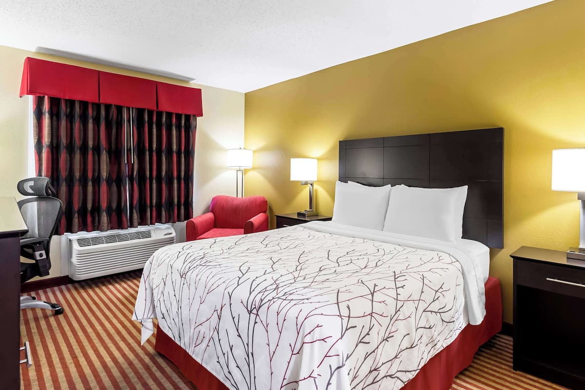 A hotel room with a large bed, a red armchair, two bedside lamps, a desk with a chair, and an air conditioning unit.