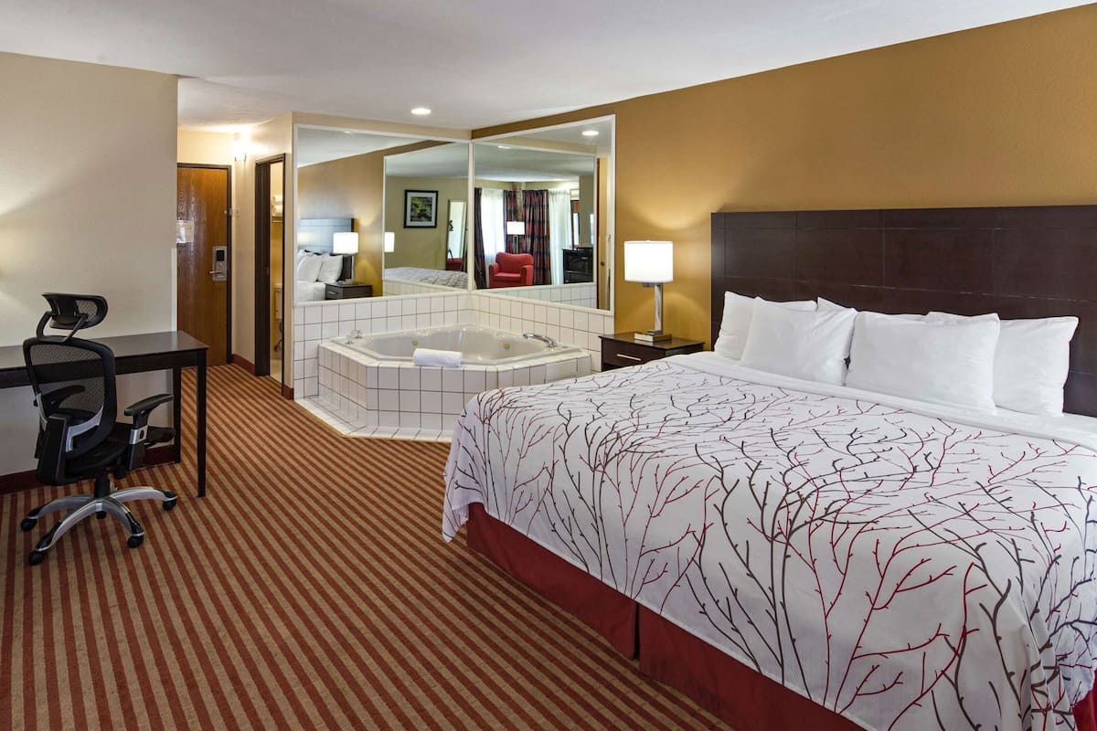 A hotel room with a bed, a desk with a chair, a desk lamp, a large mirror, a bathtub, and a carpeted floor.