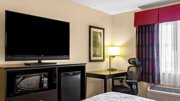 A hotel room with a flat-screen TV, a microwave, a mini-fridge, a desk with a chair, a bed, a window with curtains, and a wall-mounted lamp.