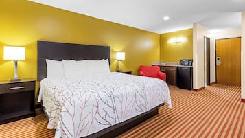 Habitación de hotel con una cama grande, dos mesitas de noche con lámparas, una silla roja, una pequeña cocina americana y una puerta de madera.