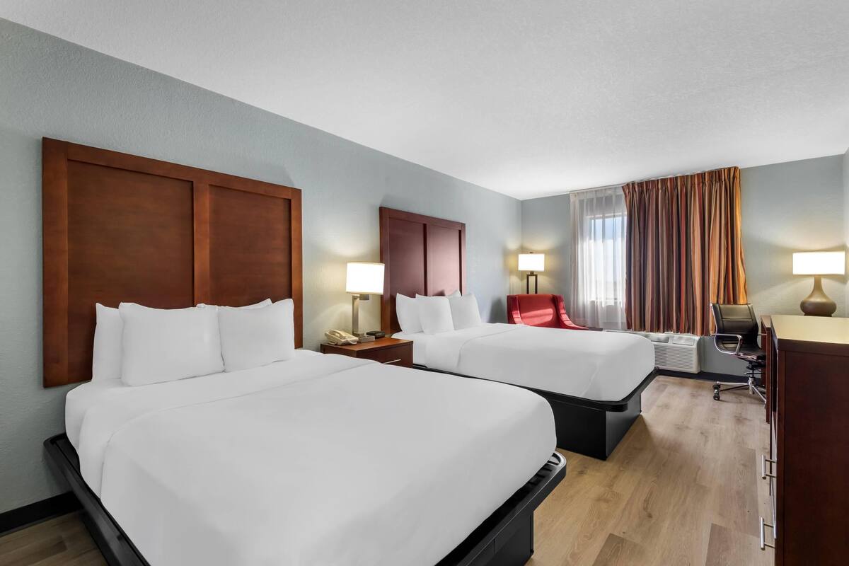 Quarto de hotel com duas camas, uma escrivaninha, uma cadeira, um abajur e uma janela com cortinas.