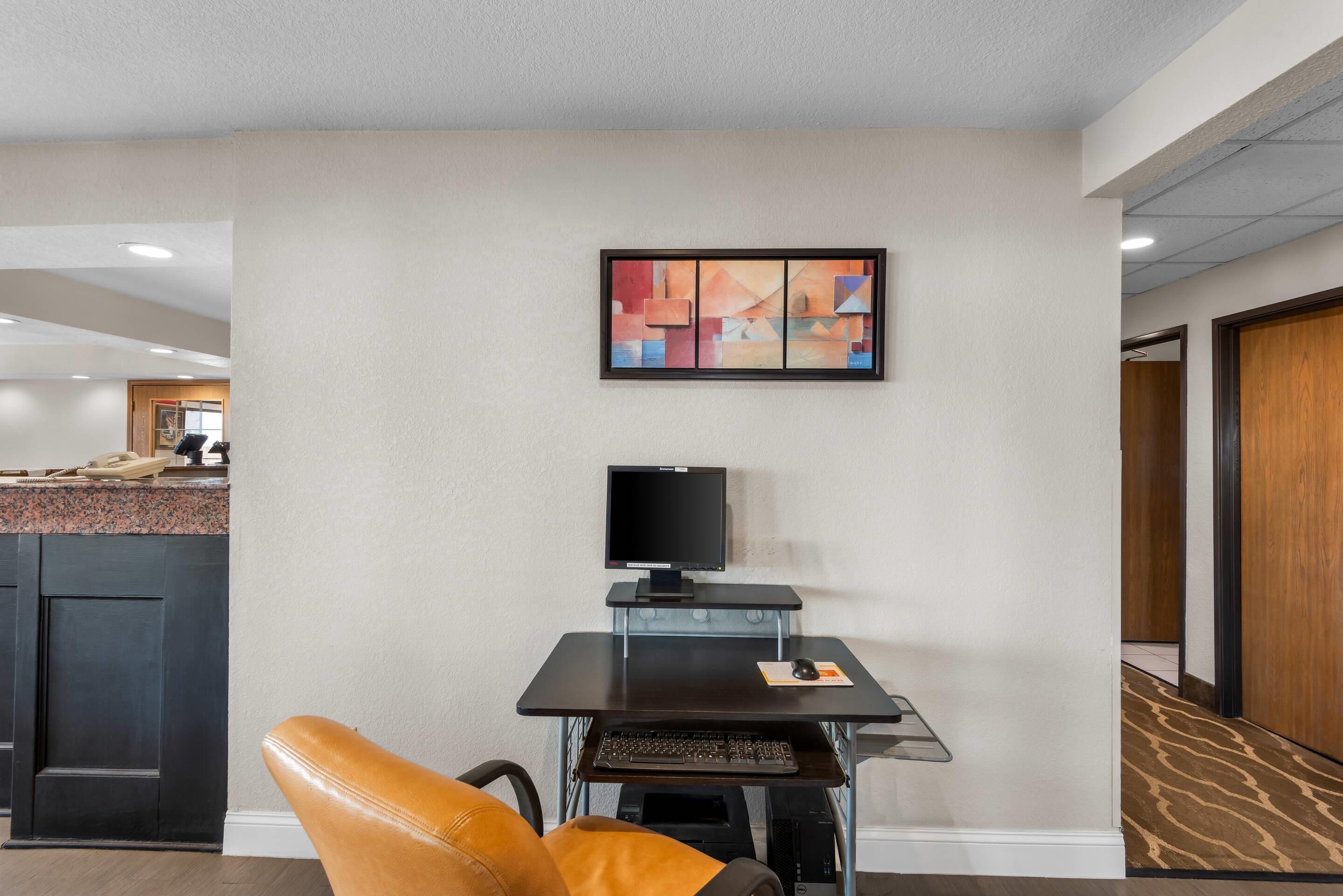 A hotel room with a desk, chair, computer, and artwork on the wall.