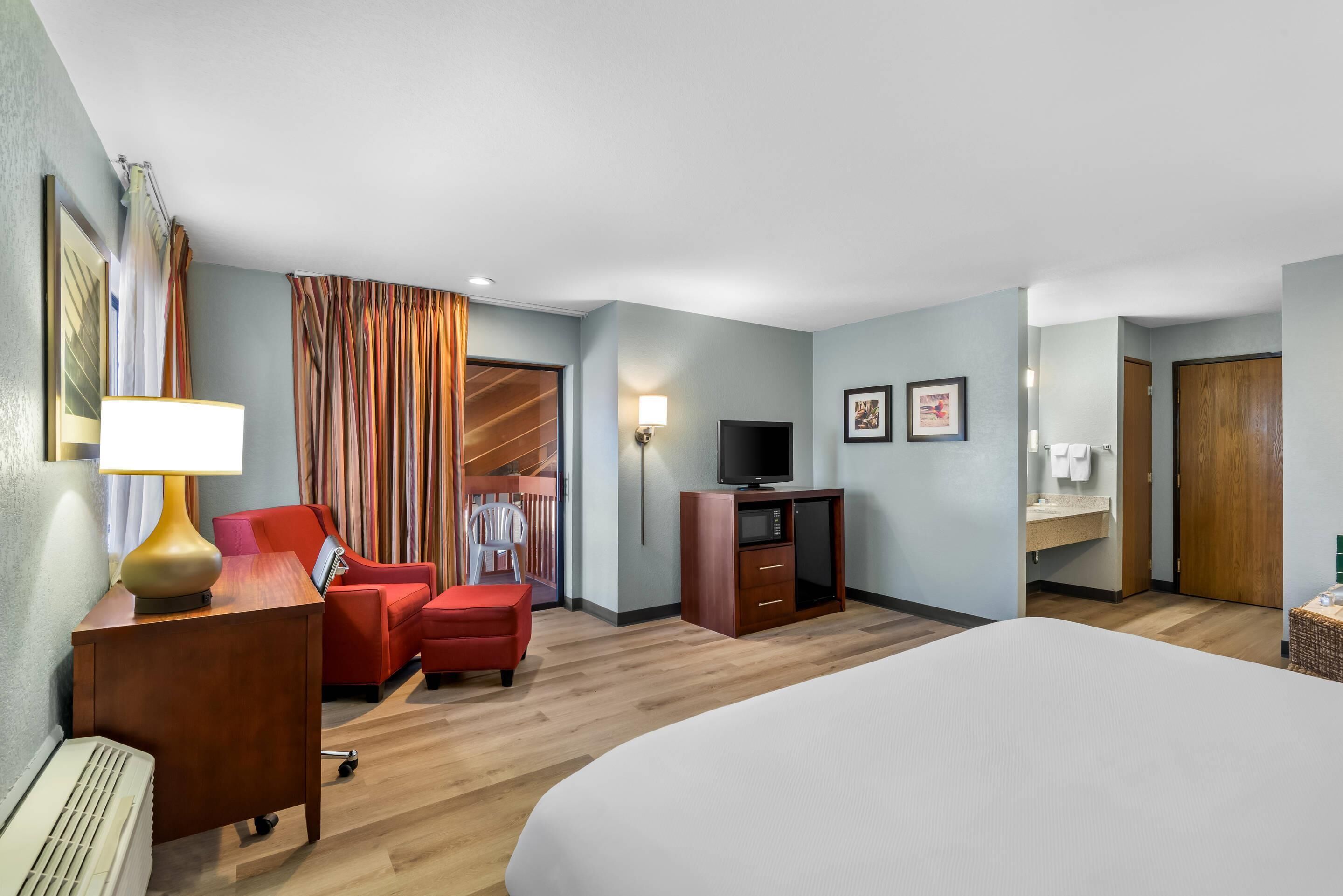 A hotel room with a bed, a desk with a lamp, a TV, a red chair, a wooden wardrobe, and a bathroom visible through an open door.