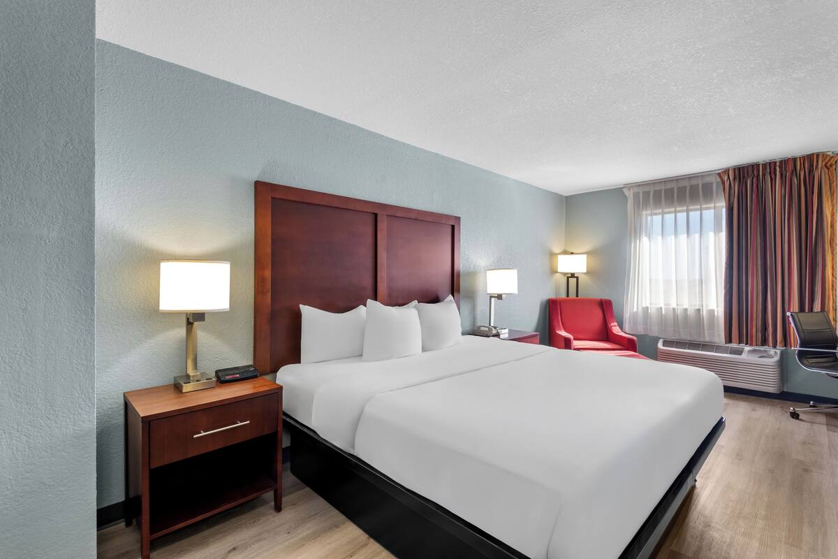 Quarto de hotel com uma cama grande, mesas de cabeceira com luminárias, uma cadeira vermelha e uma janela com cortinas.