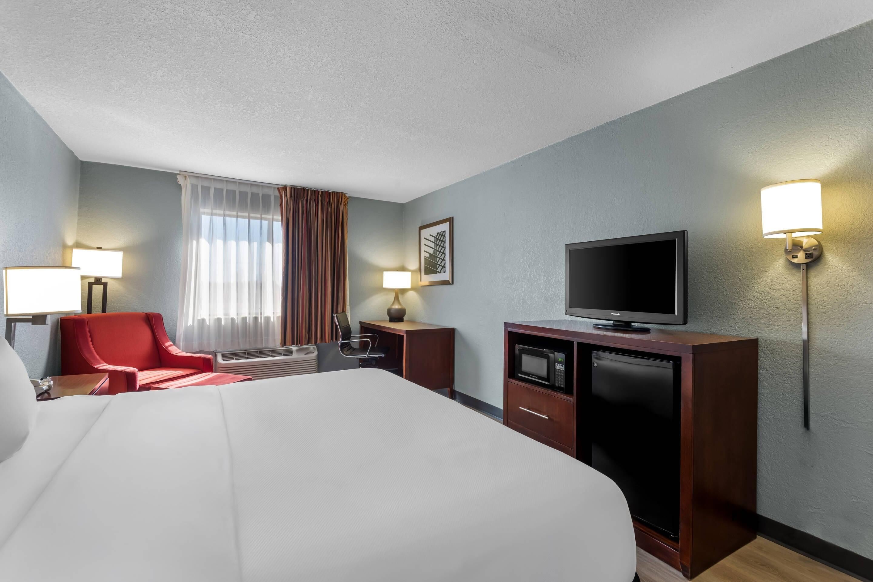 Ein Hotelzimmer mit einem großen Bett, einem Fernseher, einem Schreibtisch mit Lampe, einem roten Sessel und einem Fenster mit Vorhängen.