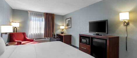 Ein Hotelzimmer mit einem großen Bett, einem Fernseher, einem Schreibtisch mit Lampe, einem roten Sessel und einem Fenster mit Vorhängen.