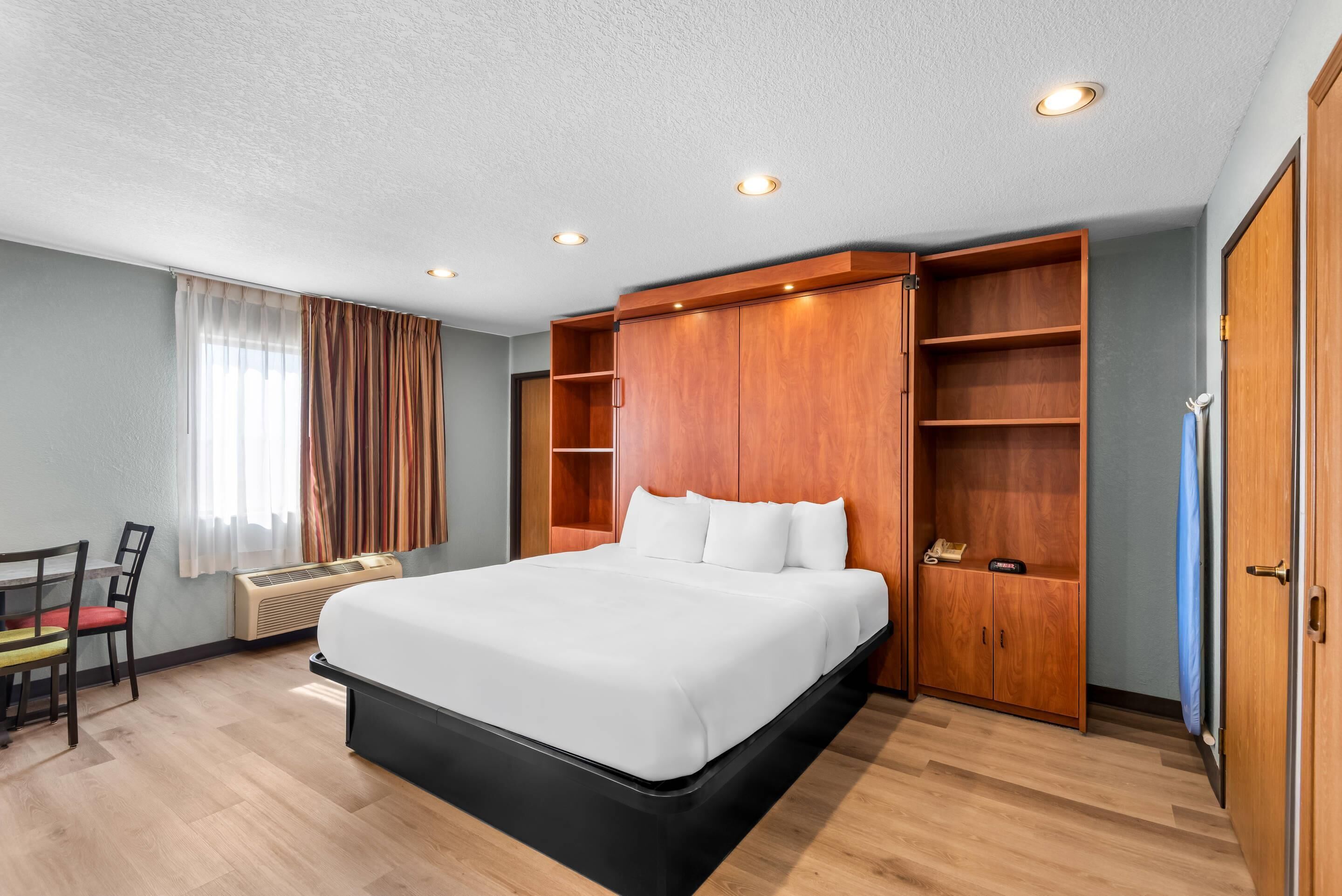 A hotel room with a large bed, wooden wardrobe, a desk with a chair, and a window with curtains.