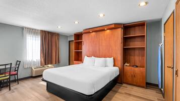 A hotel room with a large bed, wooden wardrobe, a desk with a chair, and a window with curtains.