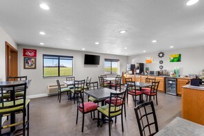 A spacious dining area with multiple tables and chairs, a kitchen area with modern appliances, and large windows offering a view of the outdoors.