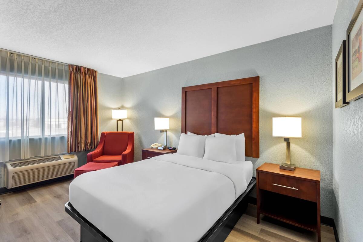 Quarto de hotel com uma cama grande, duas mesas de cabeceira com luminárias, uma cadeira vermelha e uma janela com cortinas.