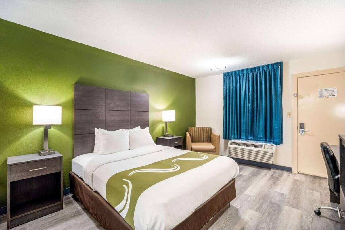 A hotel room with a large bed, two bedside tables with lamps, a chair, a desk, and a window with curtains.