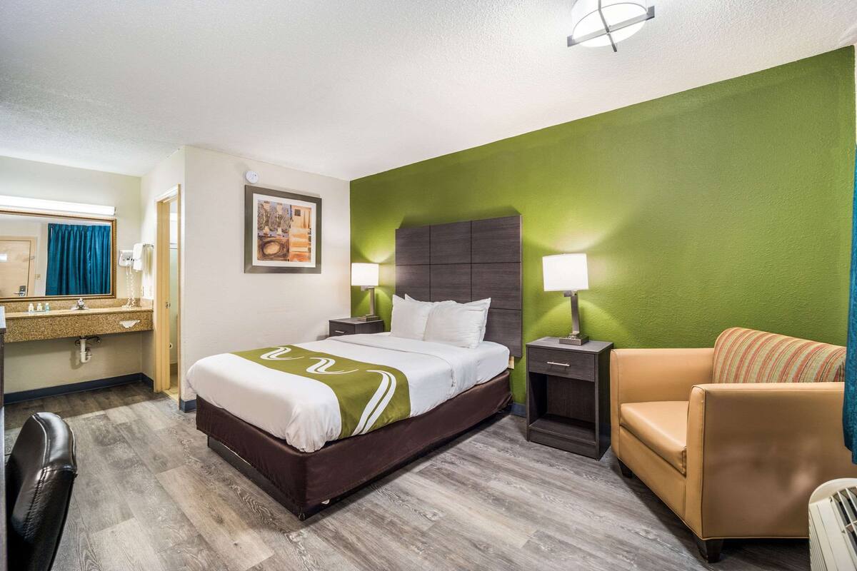 A hotel room with a bed, a chair, a desk with a mirror, and a green accent wall.