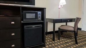 A compact hotel room with a kitchenette including a microwave, refrigerator, and a small TV. There's a desk with a chair and a wall-mounted lamp. The room has a patterned carpet and a door in the background. - Super 8 by Wyndham Decatur/Lithonia/Atl Area (Decatur)