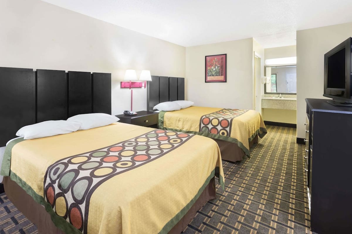 A hotel room with two beds, a desk, a TV, and a bathroom visible in the background.