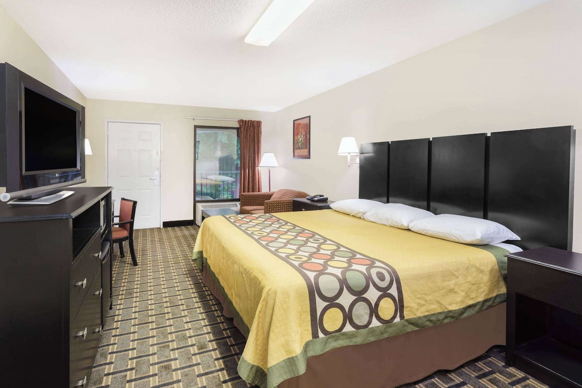 A hotel room with a bed, a desk with a TV, a chair, a nightstand, and a window with curtains.