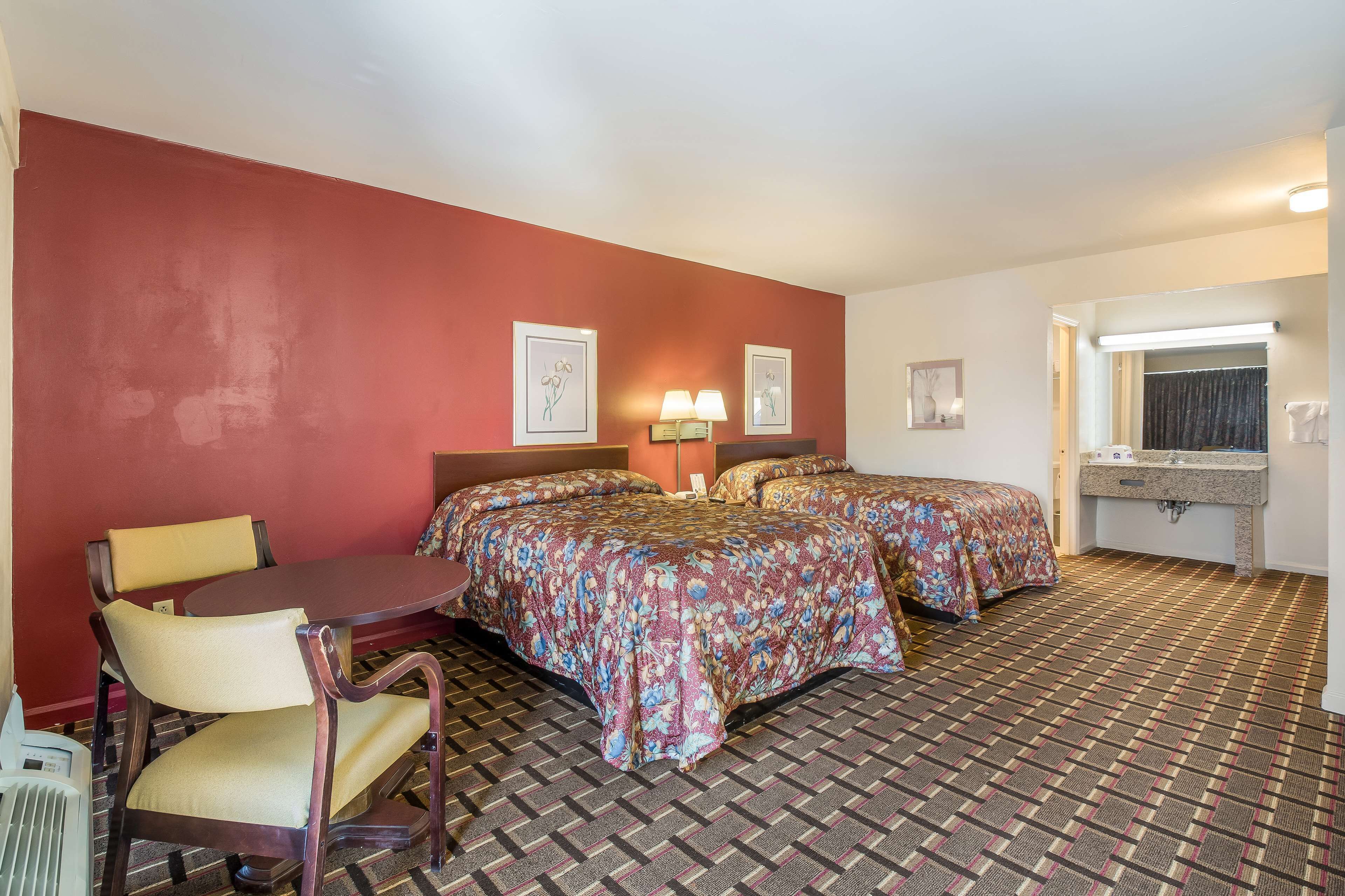 Une chambre d’hôtel avec deux lits, un bureau, une chaise, une lampe de bureau, un miroir et une salle de bain équipée d’un lavabo et d’un porte-serviettes.