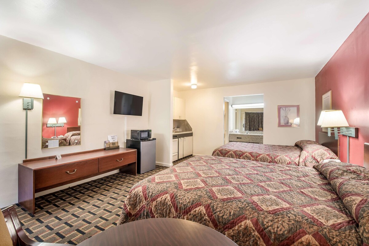 A hotel room with two beds, a desk, a TV, a kitchenette, and a bathroom.