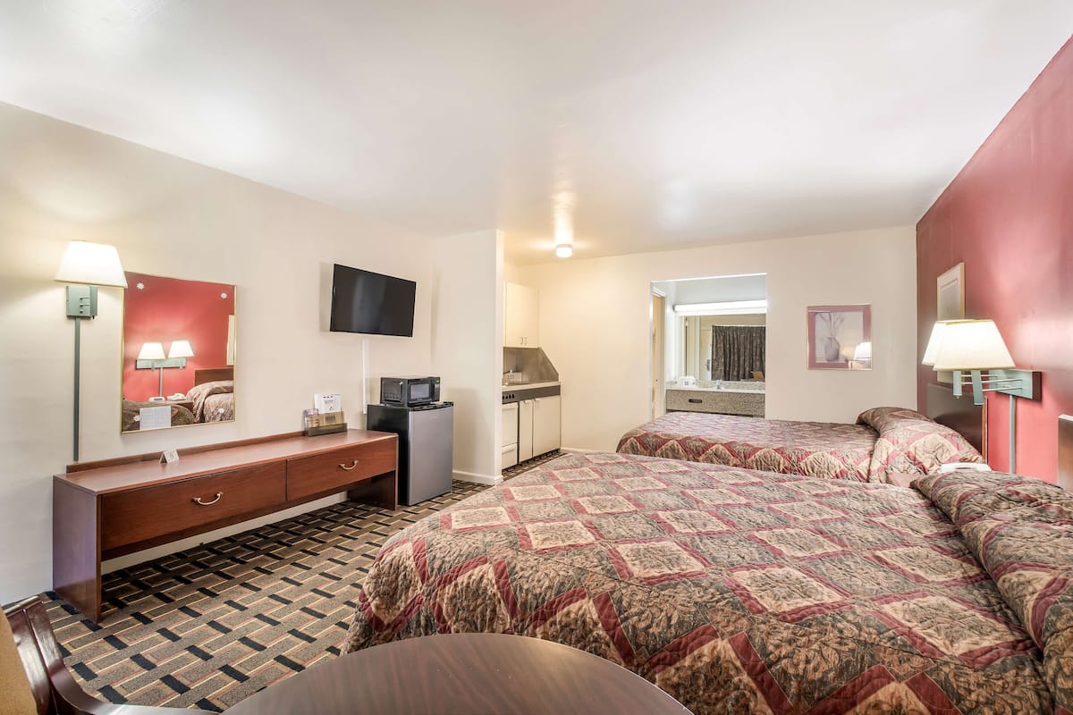 Habitación de hotel con dos camas, un escritorio, televisor, kitchenette y un baño visible a través de una puerta abierta.
