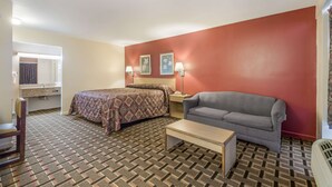 A room with a bed, a gray sofa, a small table, a nightstand with a lamp, and a window with a curtain. - Knights Inn Cleveland, TN (Cleveland)