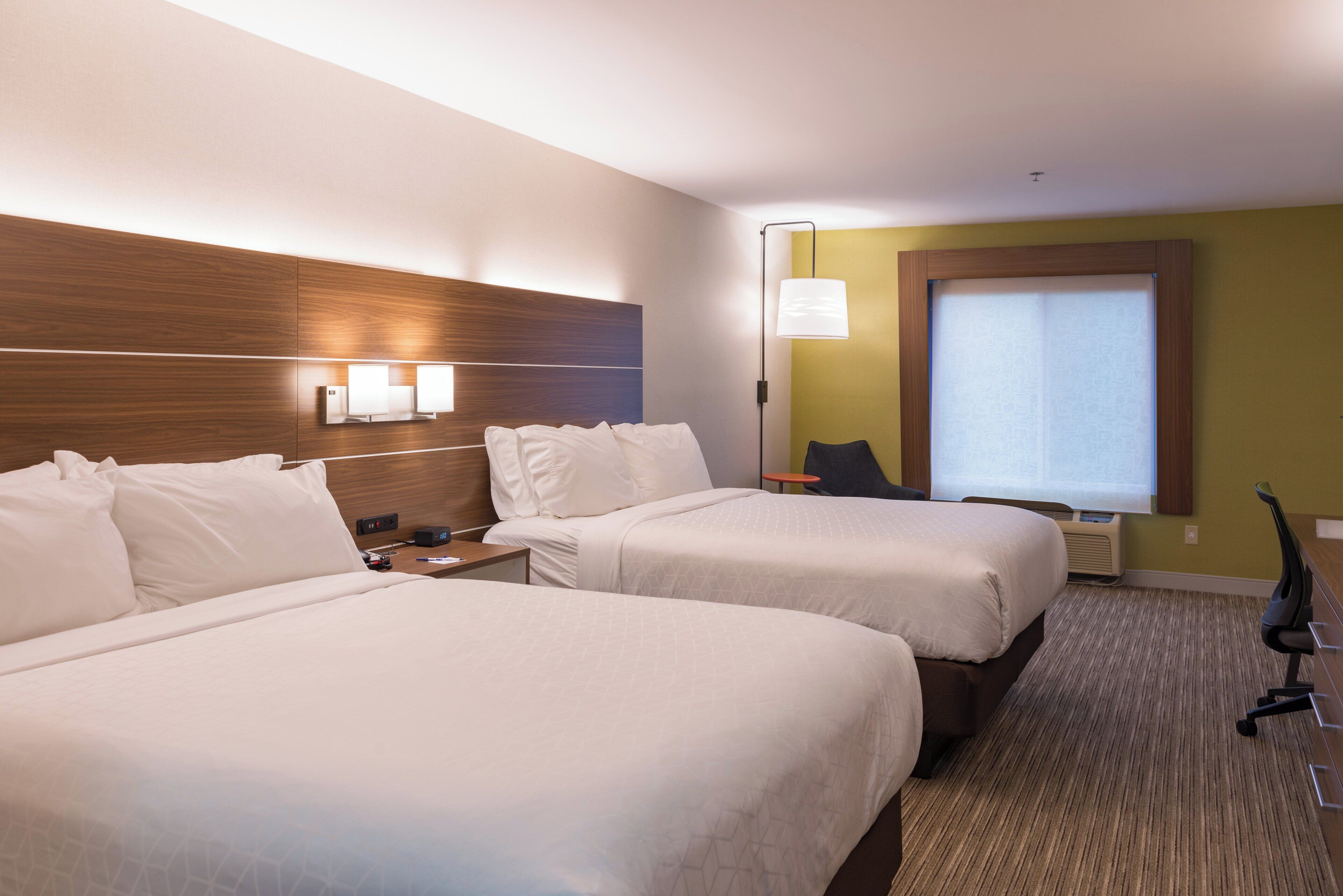 A hotel room with two beds, a nightstand, a lamp, a chair, and a window with blinds.