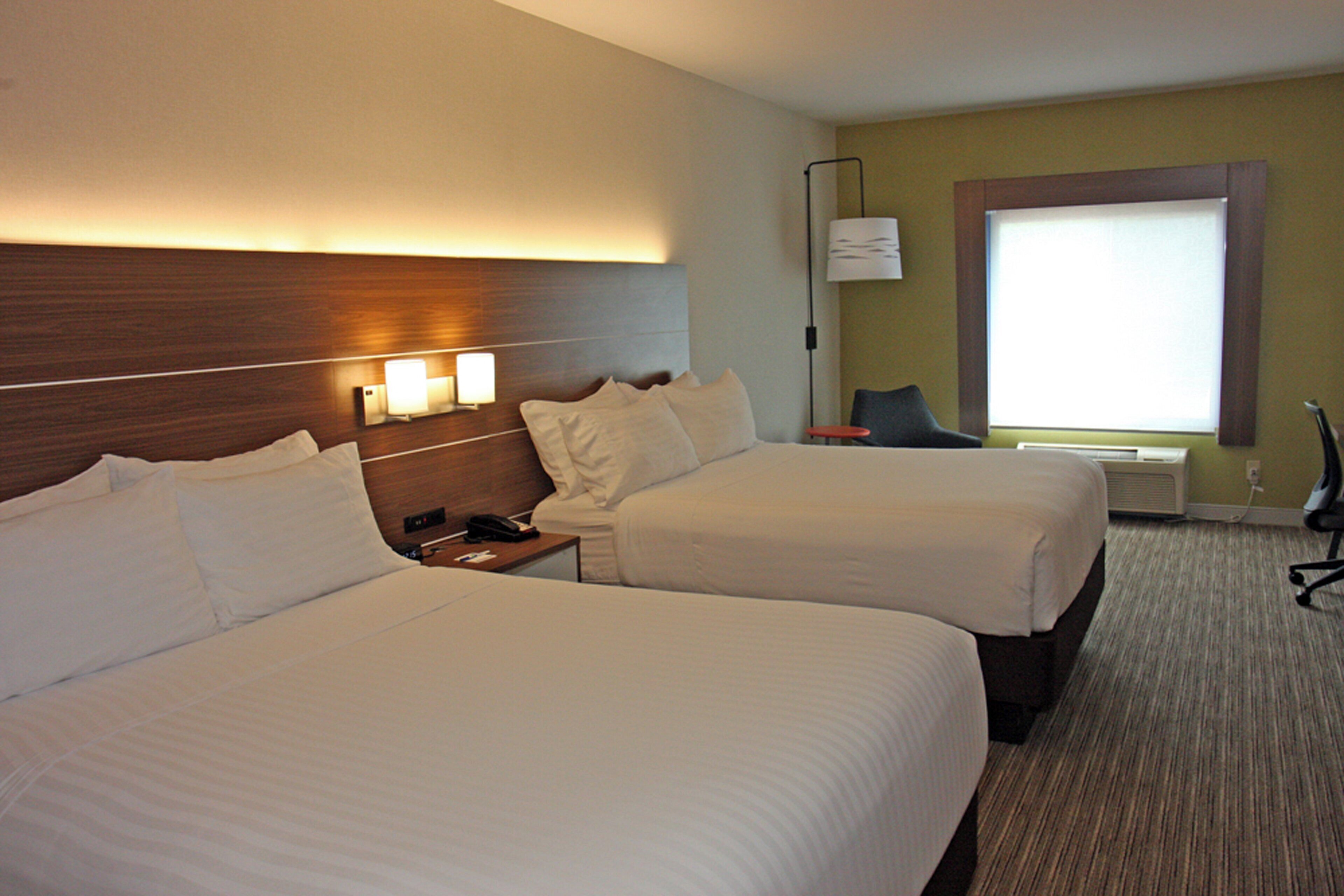 A hotel room with two beds, a nightstand, a lamp, a chair, and a window with blinds.
