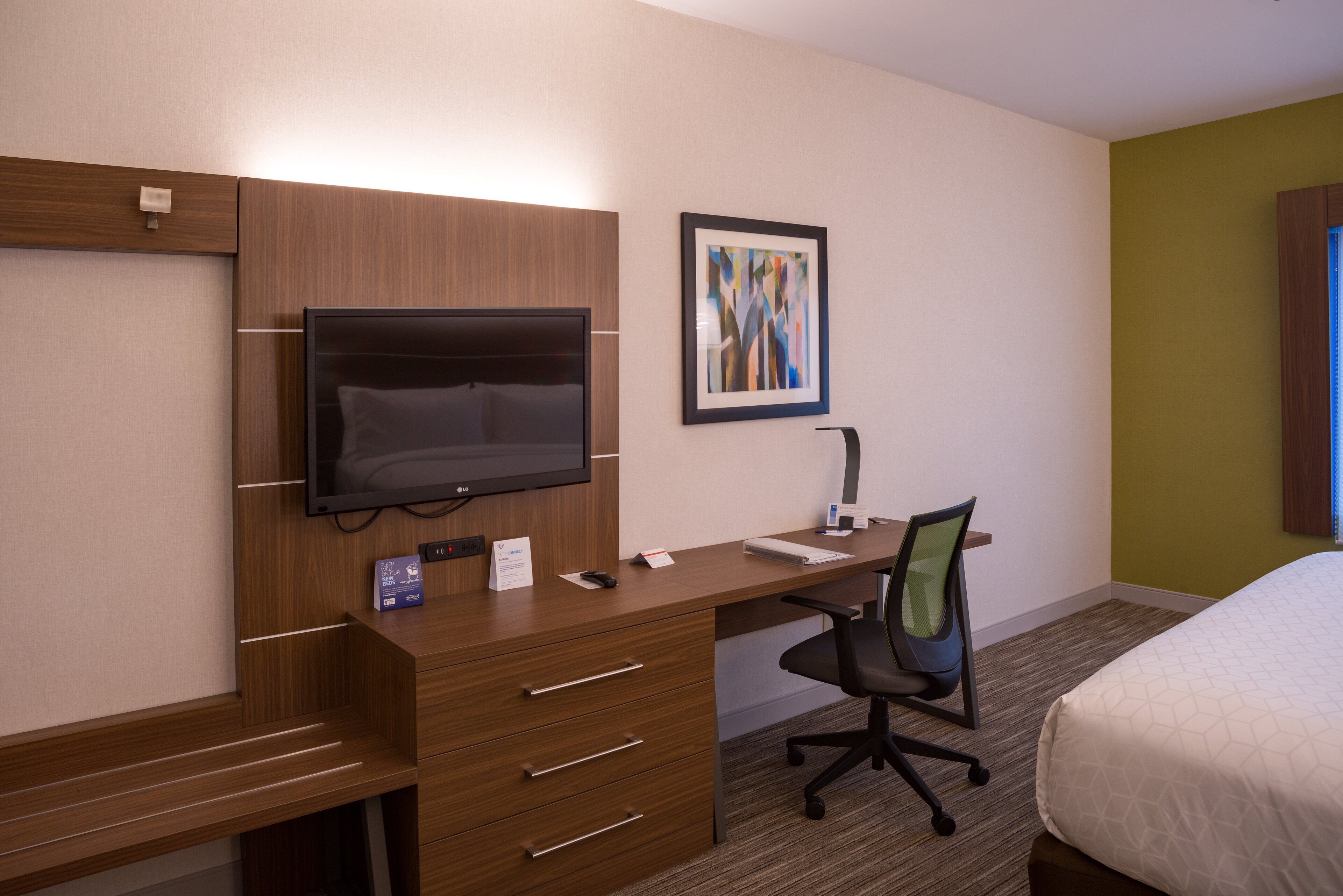 Quarto de hotel com secretária de madeira, televisão de tela plana, cadeira, cama e pintura abstrata emoldurada na parede.