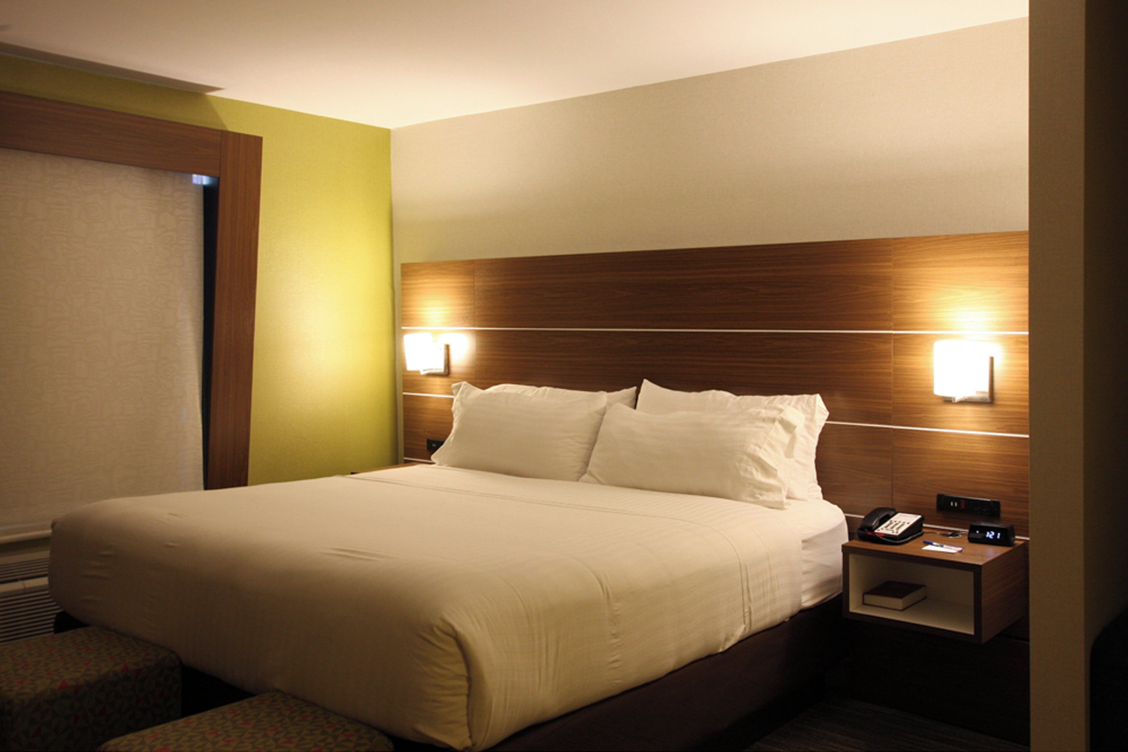 Une chambre d’hôtel avec un grand lit, des lampes de chevet, une table de chevet avec un téléphone et une horloge, et une fenêtre avec des stores.