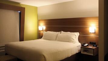 Une chambre d’hôtel avec un grand lit, des lampes de chevet, une table de chevet avec un téléphone et une horloge, et une fenêtre avec des stores.
