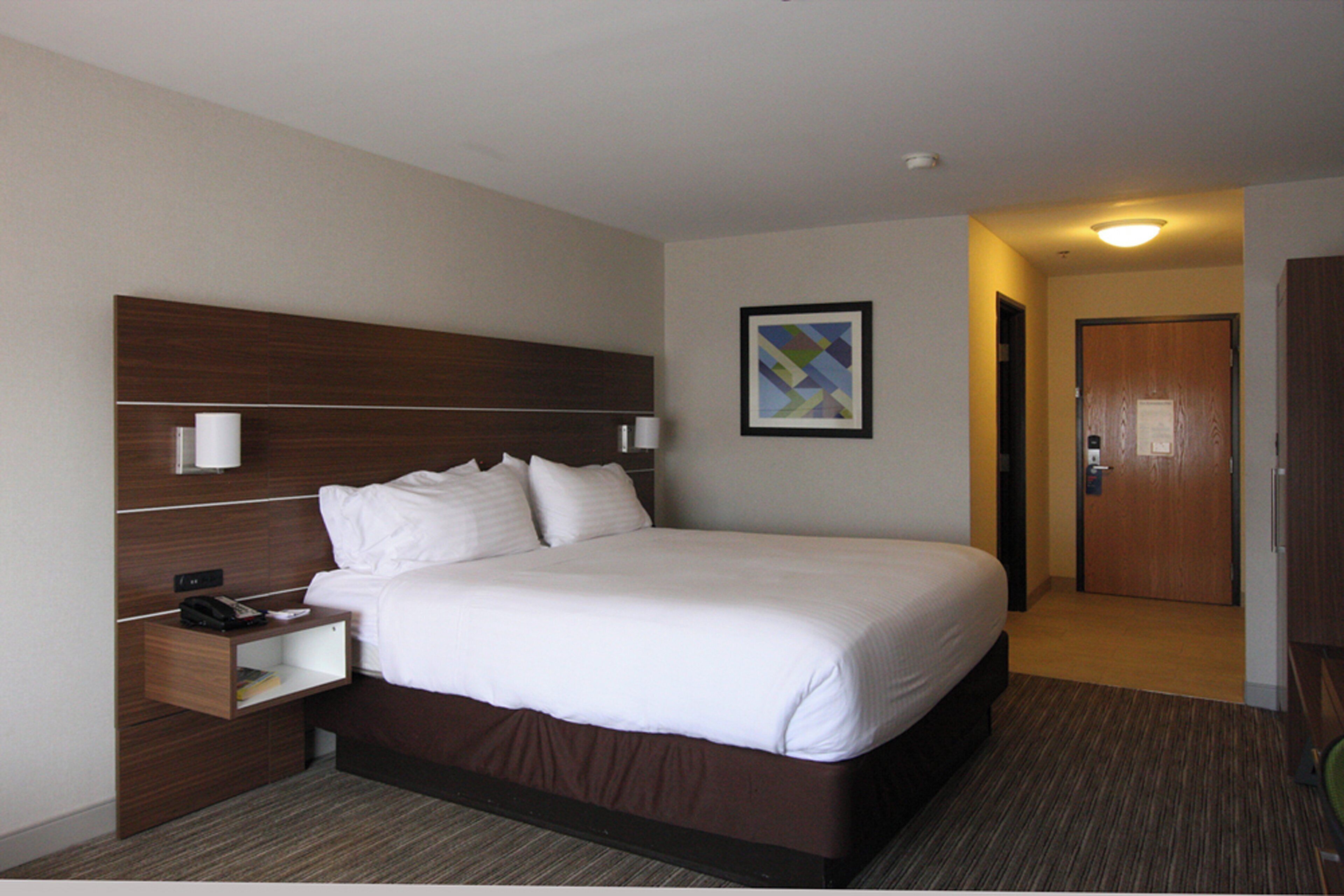 A hotel room with a large bed, a bedside table with a lamp, a nightstand, a framed picture on the wall, and a door leading to another room.