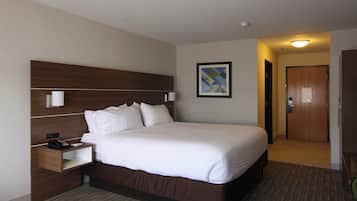 A hotel room with a large bed, a bedside table with a lamp, a nightstand, a framed picture on the wall, and a door leading to another room.