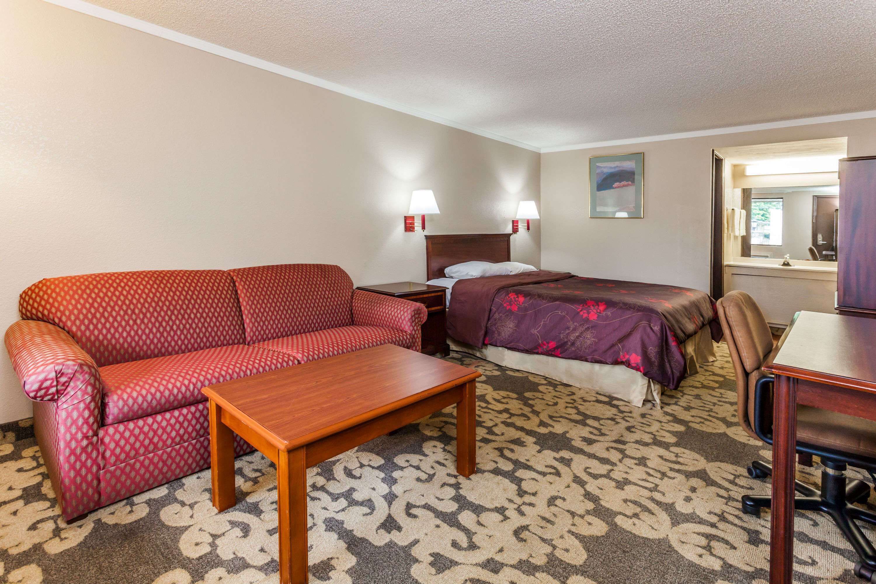 Quarto de hotel com um sofá vermelho estampado, uma mesa de madeira, uma cama com colcha florida roxa e uma escrivaninha com uma cadeira.