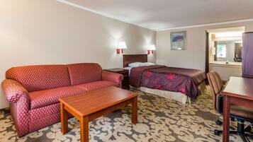 Quarto de hotel com um sofá vermelho estampado, uma mesa de madeira, uma cama com colcha florida roxa e uma escrivaninha com uma cadeira.