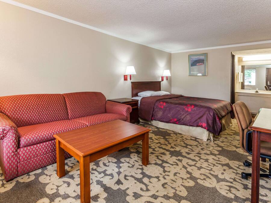 Quarto de hotel com um sofá vermelho estampado, uma mesa de madeira, uma cama com colcha florida roxa e uma escrivaninha com uma cadeira.