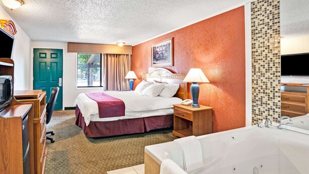 Habitación de hotel con cama, escritorio, silla, lámpara de escritorio, mesita de noche, teléfono, televisor, ventana con cortinas, puerta y baño con bañera.
