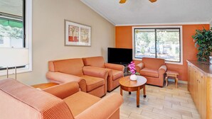 Una sala de estar con muebles naranja, un televisor de pantalla plana, una mesita con un jarrón y una ventana con vistas al exterior.