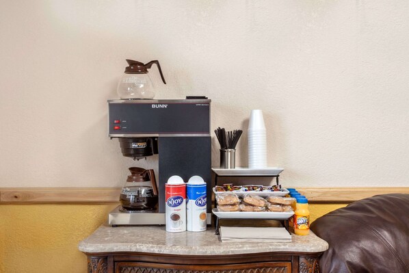 Uma estação de café com uma máquina de café Bunn, dosadores de açúcar e creme, copos descartáveis, uma bandeja de pastelaria e um pote de mel.