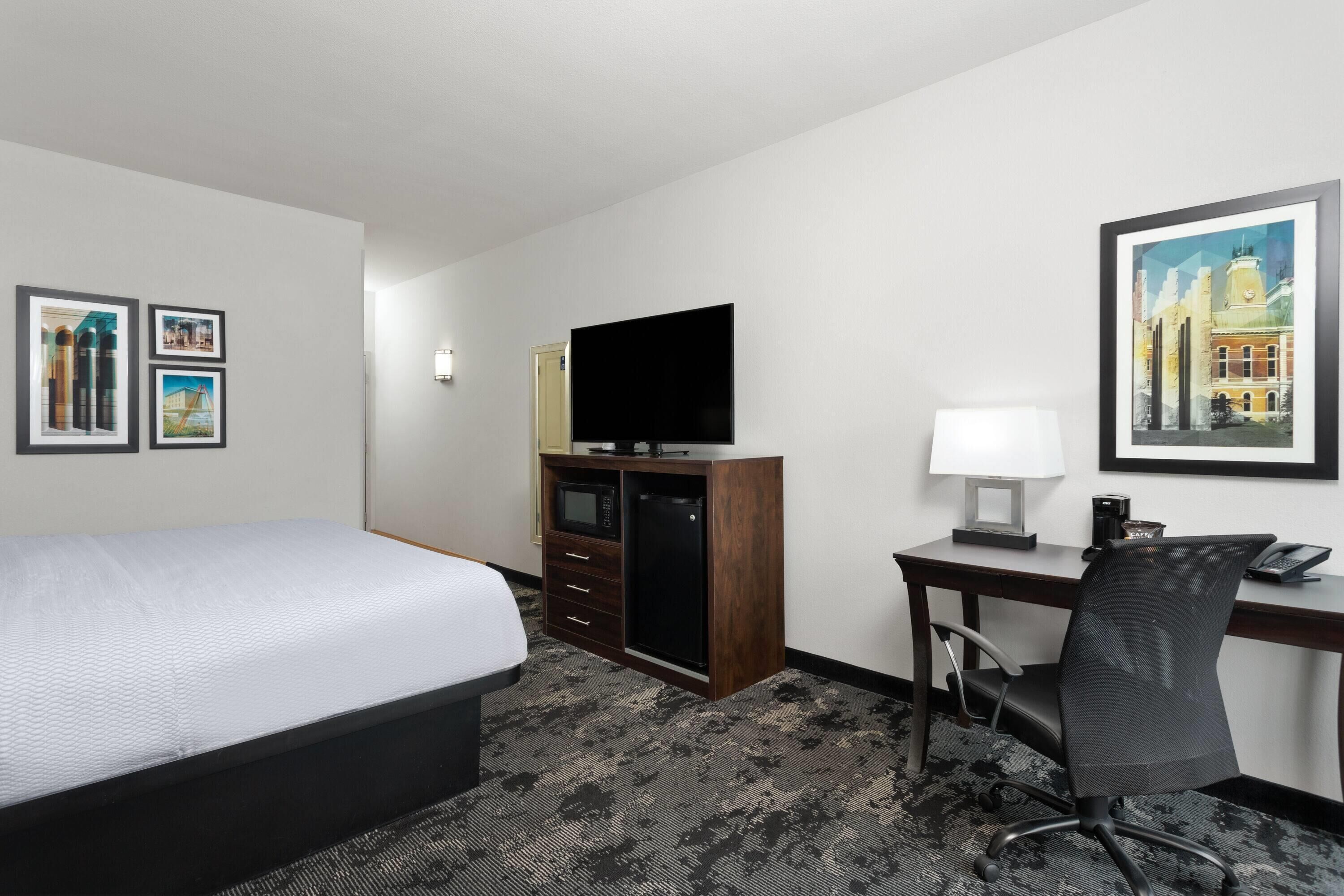 A hotel room with a bed, a desk with a computer, a TV, and framed pictures on the walls.