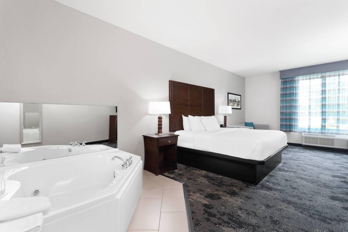 A modern hotel room with a large bed, a separate bathroom with a bathtub, and a large window with curtains.