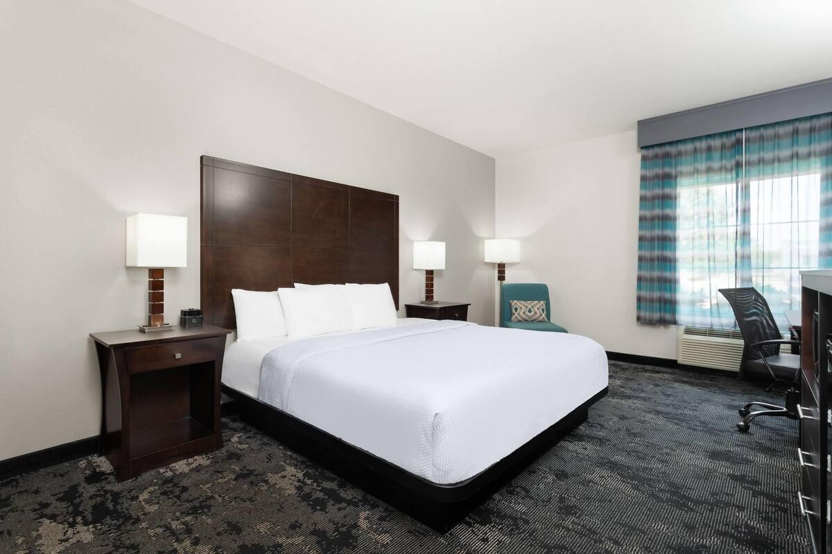 A hotel room with a large bed, two bedside tables with lamps, a desk with a chair, and a window with curtains.