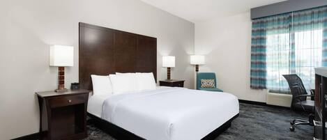 Quarto de hotel com uma cama grande, duas mesas de cabeceira com luminárias, uma escrivaninha com cadeira e uma janela com cortinas.