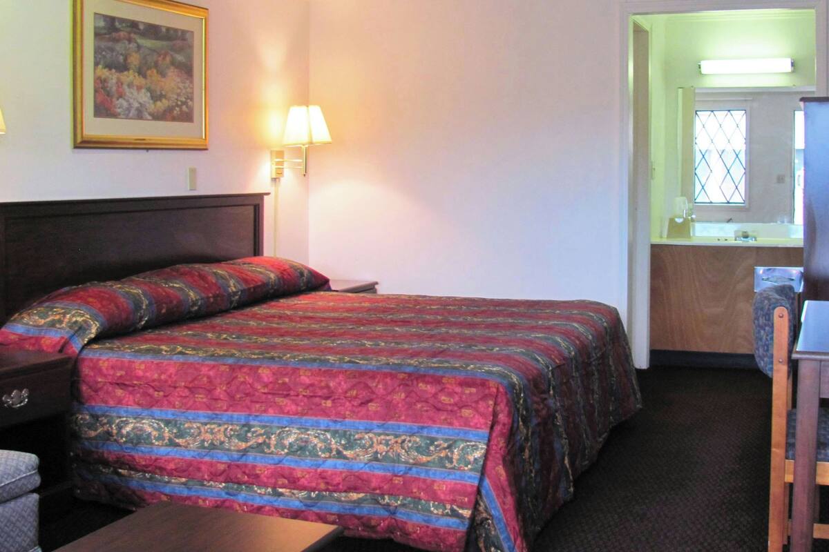 A hotel room with a bed, a desk, a chair, a lamp, a painting, and a window.