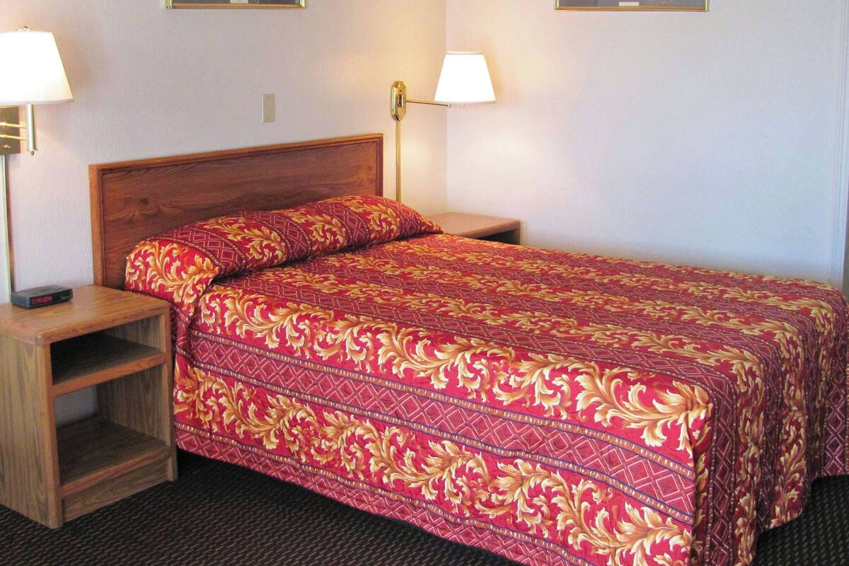 A hotel room with a wooden headboard, a red patterned bedspread, two bedside lamps, and a wooden nightstand with a shelf.